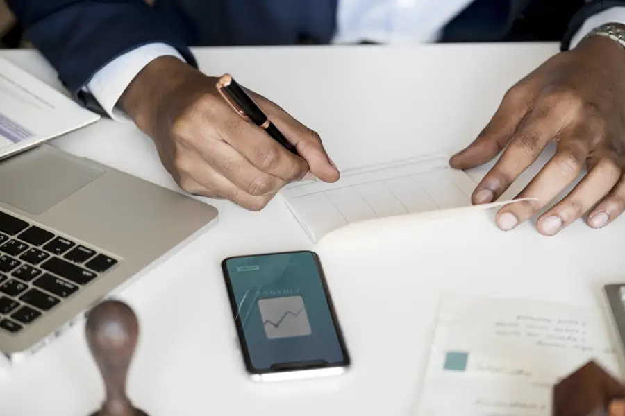 Hands signing a document beside a phone and a laptop